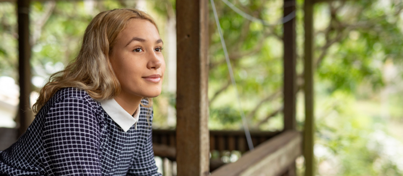 A young woman stands on her porch, looking at the trees. She has started mindfulness practices and it has supported her mental health.