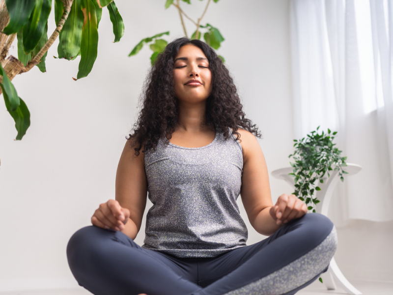 A young woman sits on the floor of her house in athletic clothes. She is a beginner to mindfulness practices.