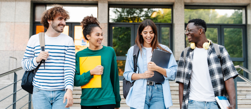 Four friends are walking together away from their class. They are notebooks, computers, and bookbags. They have just started school again for the Fall.