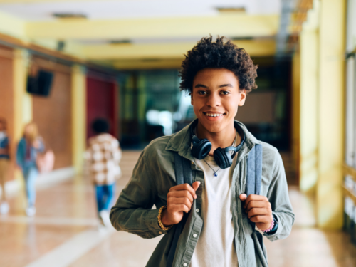 A male high school student in the hallway during his first day of classes for the Fall. He is wearing a tan shirt with a green jacket and carrying his bookbag.