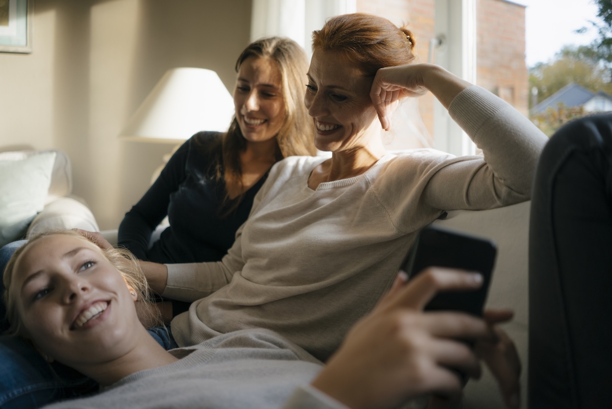 Happy family of mother and two daughters, spending time together smiling, while one daughter is using social media.