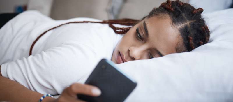 Young woman laying in bed on her phone in a white shirt. The woman experiences a trauma response called fawning.