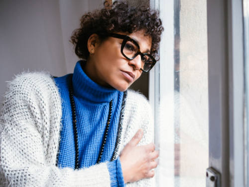 A young woman stands in her house, looking at the snowfall. She is feeling the holiday blues this winter.