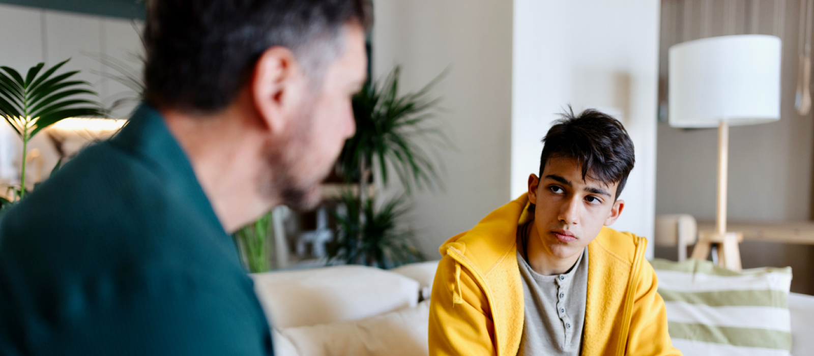 A young male talks with his dad on the couch. He is talking through his triggers to prevent a self-harm relapse.