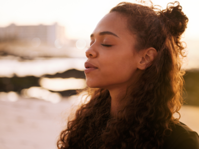 A young Black woman struggling with mental health stands with her eyes closed at sunset