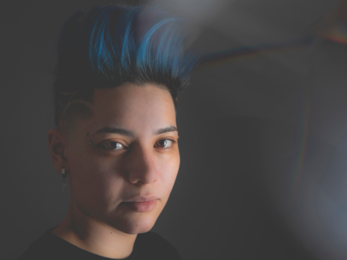 An LGBTQIA+ teen with blue hair looks directly at the camera