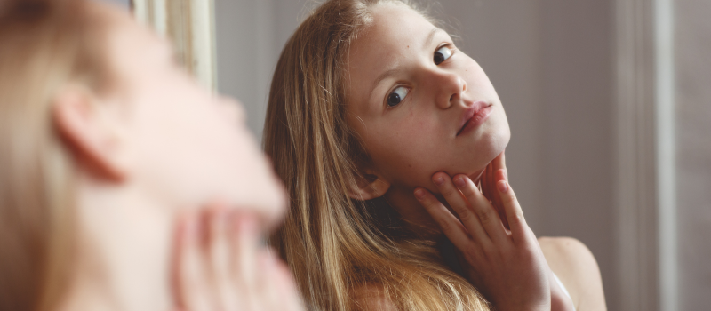 A young teen girl with blonde hair stares at herself in the mirror