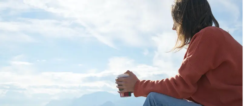 A young woman holding a cup of coffee sits alone outside staring 