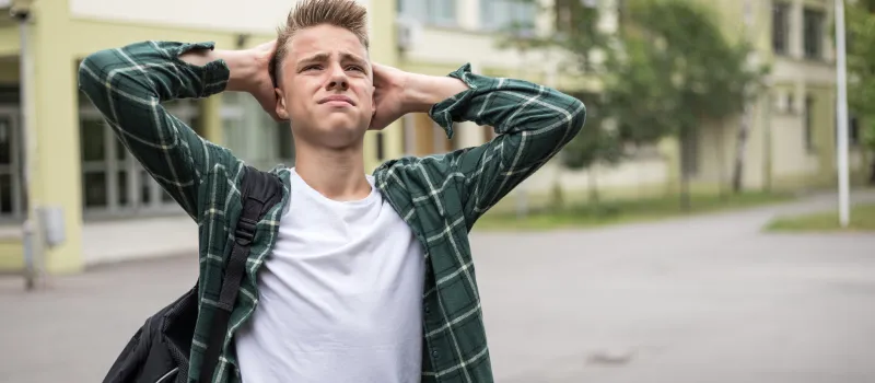 A young man with blonde hair in a green plaid shirt looks distressed with his hands behind his head