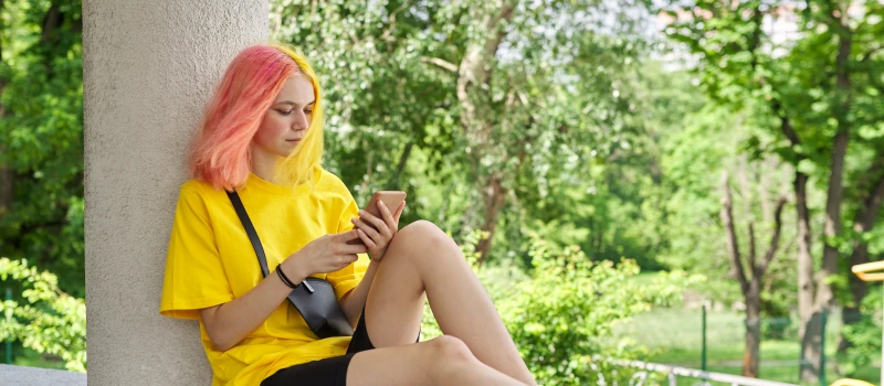 A teen girl in a yellow tshirt with pink and yellow hair sits outside on her phone with a fanny pack
