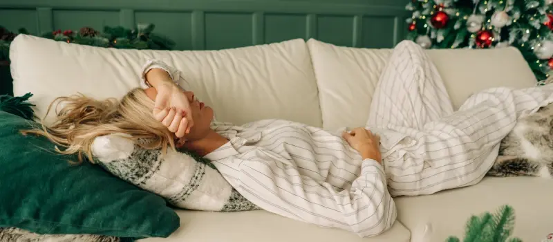 A woman lays on her couch in pajamas, sad at Christmas time