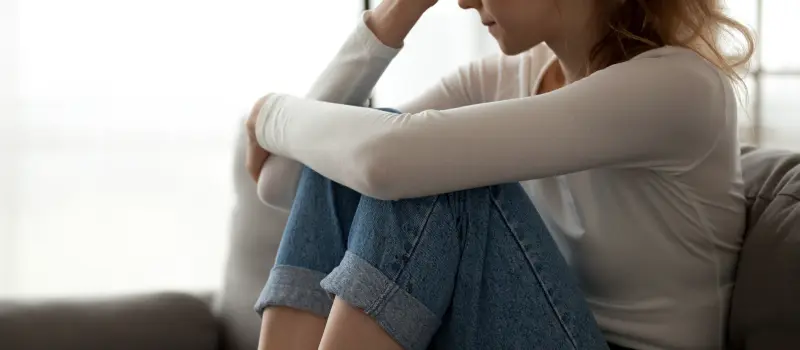 A teen girl in a white shirt sits on a couch hugging her knees into her chest looking sad.