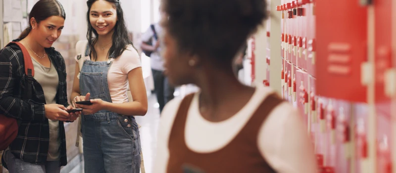 A teen girl standing in front of her locker looks back at two other teens who are laughing at her.