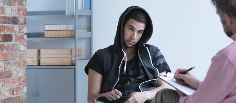 A teen boy in a sweatshirt sits with his hood up staring off into space during therapy