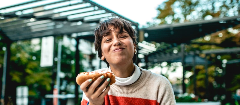 A teen woman with short brown hair smiles at a camera holding a hot dog