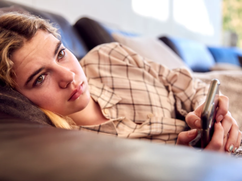 A young woman with blonde hair lays on her couch holding her cell phone.