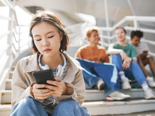 A teen girl with short hair sits on the steps at school on her phone while a group of teens behind her talks