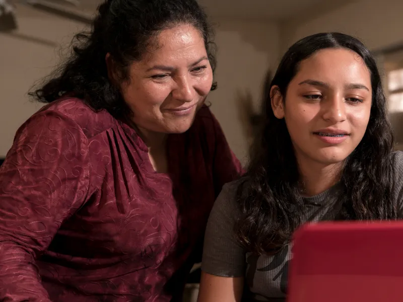A mom in a red shirt sits with her daughter in a black shirt on a red laptop during virtual family therapy