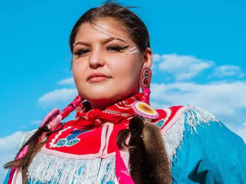An Indigenous woman in traditional Native American clothing and makeup stares directly at the camera.