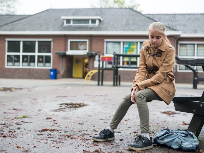 A young girl with blonde hair in a tan coat sits alone in a school yard