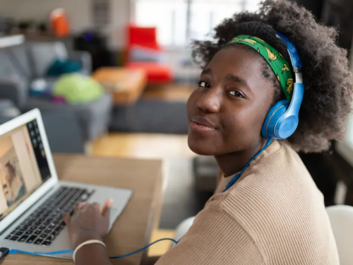 A teen girl with curly hair sits at her laptop wearing bright blue headphones