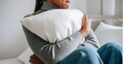 A teen in a gray sweater and jeans sits on her bed hugging a pillow.