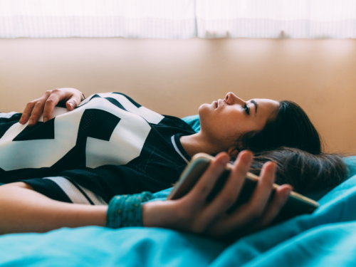 A teen with brown hair lays in bed holding her cell phone and stares at the ceiling