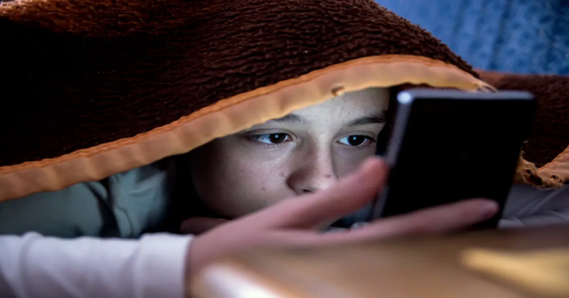 A young male teen being cyberbullied under a blanket at home.