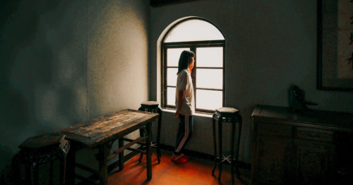 A young woman stands in front of a window looking outside as the season changes