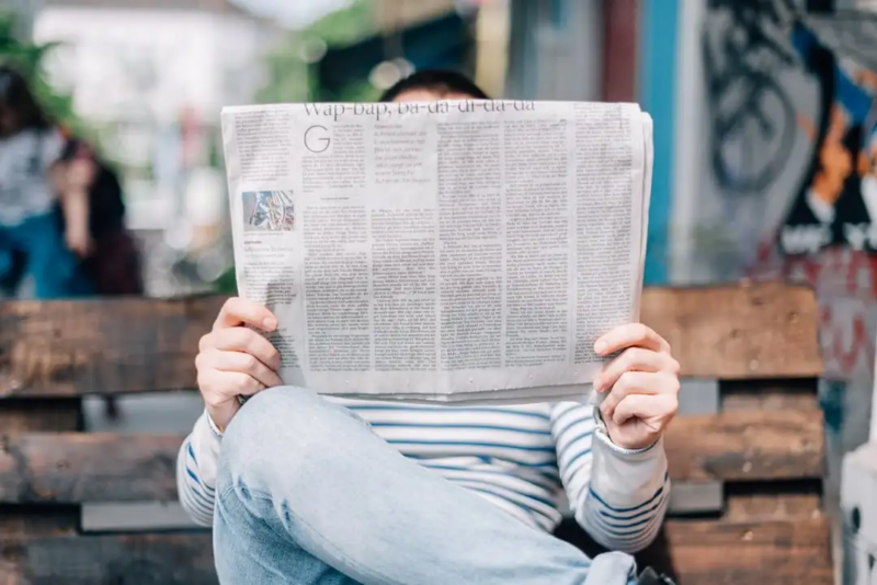 A person sits on a bench reading a newspaper that is open and covering their face