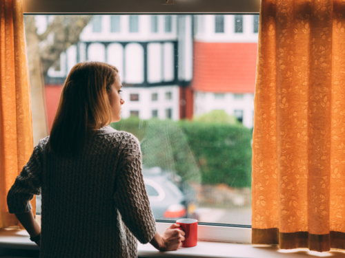 A young woman struggling with mental health issues stands at home looking out of a window
