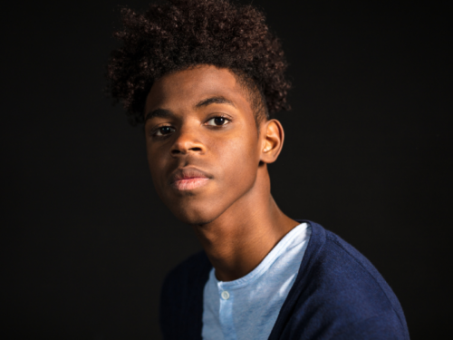 A young man wearing a blue t-shirt and navy blue cardigan staring pensively into the camera with a solid black background.