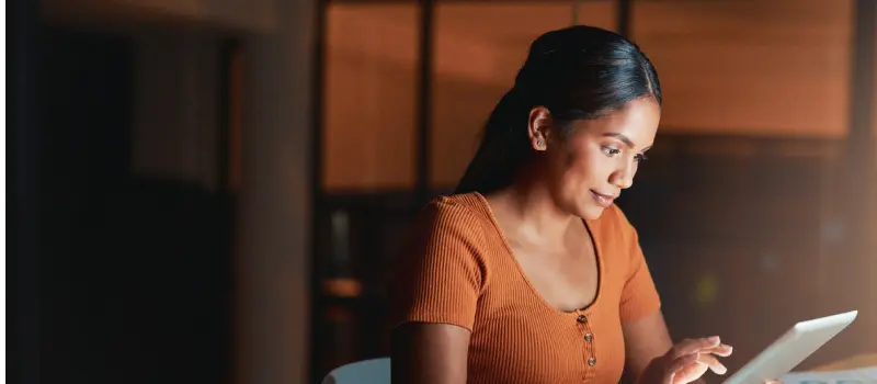 A woman sits in a dark room alone, browsing the web on a tablet.