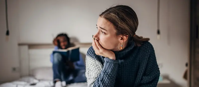 Two individuals sit on a bed together. One of them looks relaxed and is reading a book. The other looks distraught and stress.