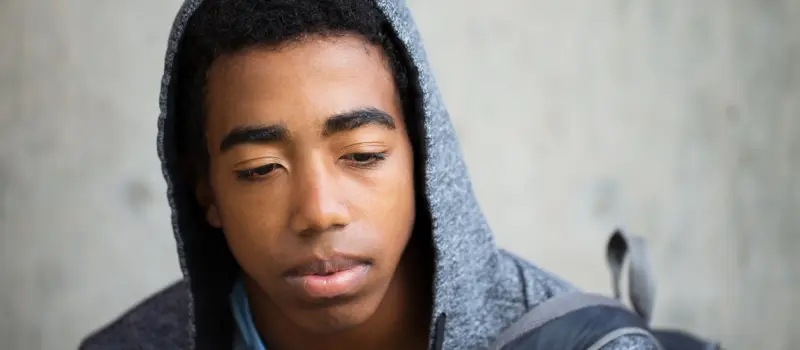 A young man coping with glass child syndrome, who is wearing a grey hoodie and a backpack, looks downward pensively.