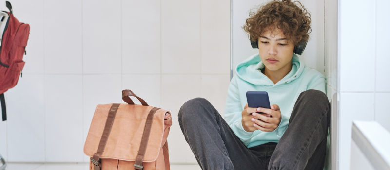 A young boy sits in a school hallway alone, looking at his phone. He appears to be anxious.