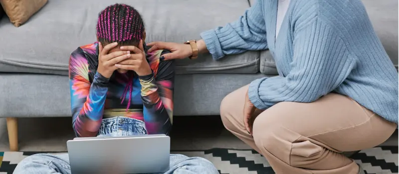 Teen dealing with anxiety holding her head while looking at a computer while her parent tries to comfort her