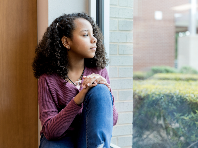 A young girl with curly hair sits by a window dealing with subconscious trauma from childhood