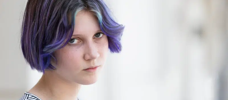 Young person with purple and blue hair dealing with trauma looks right at the camera