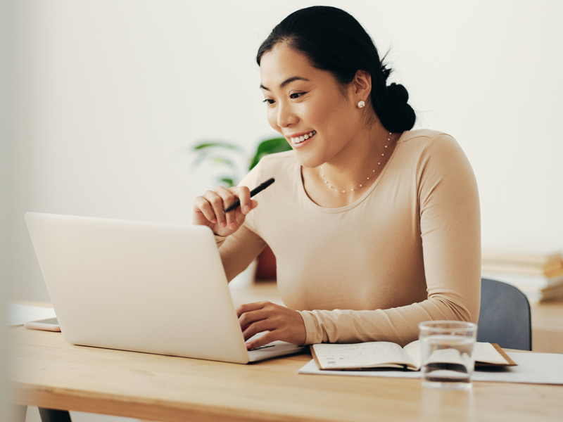 Woman with pen on laptop smiling. Notably not a mac but it looks like a mac