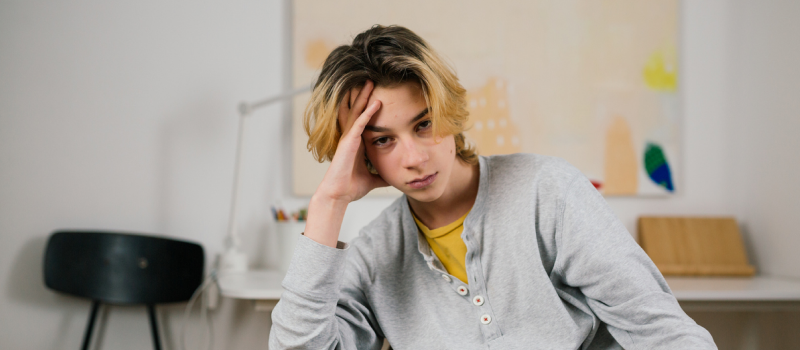 A young person with blonde hair stares into the camera with a hand on their head. Their brow is slightly furrowed from the stress of past trauma.