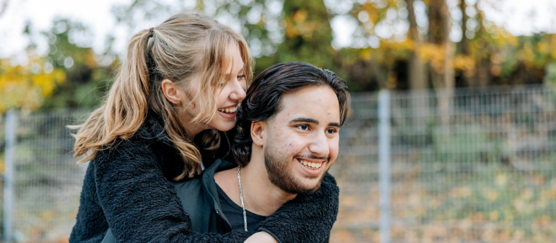 A man with a silver chain carries his girlfriend, who has OCD, on his back as both of them smile.