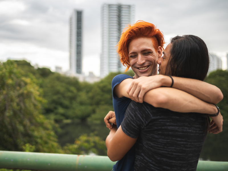Two people, one with orange hair and one with black hair, hug each other. They both are smiling and feel secure in their gender and sexuality.