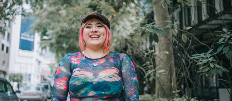 A person with pink hair and a multi-colored shirt who is secure in their gender and sexuality smiles at the camera while wearing a hat.