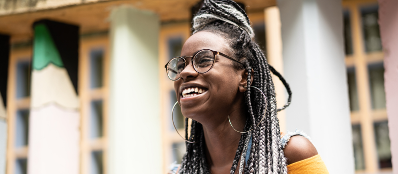 A person with braided hair and glasses smiles at the camera after learning to become more resilient.