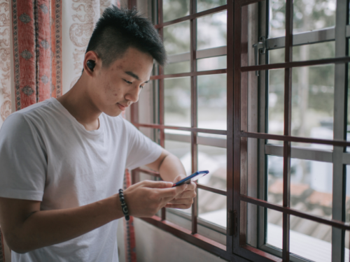 A teen in a white tshirt stands in front of a window with headphones in scrolling through TikTok