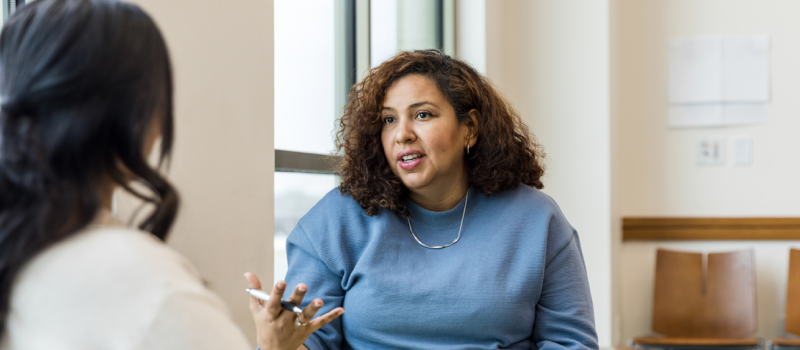 A clinician in a blue sweater explains the importance of self-care to another mental health provider during a therapy session.