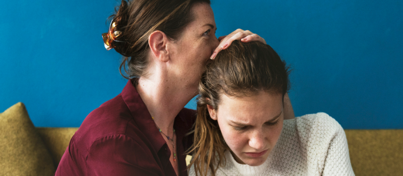 A woman in a maroon top comforts her daughter in a white sweater. The daughter has recently been treated for anxiety and suicidal ideation.