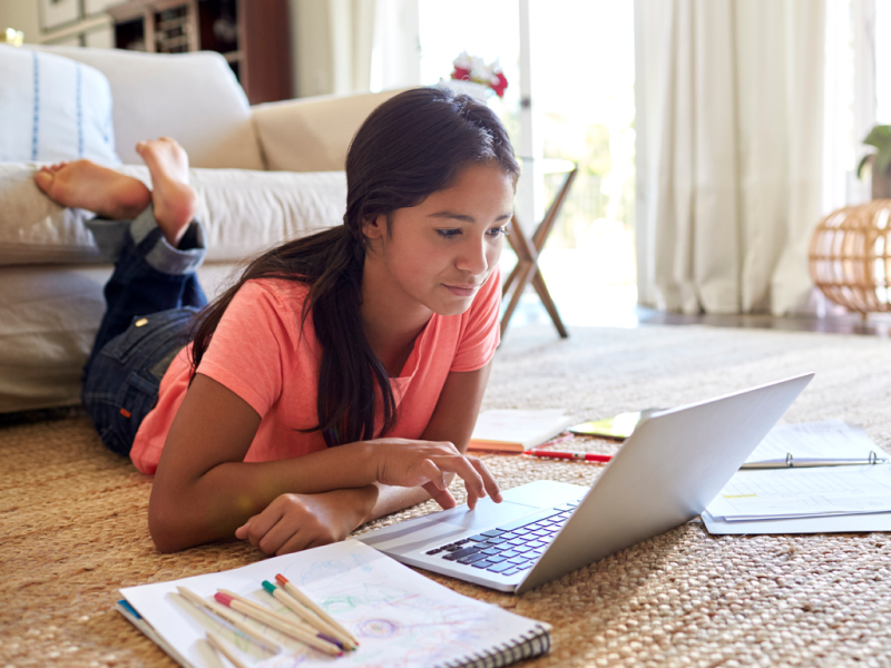 Teen laying on ground in pink shirt. She has her computer and school work out and is stimming through touch with her feet rubbing together to self-soothe her ADHD.