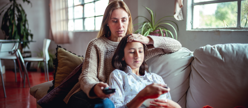 A daughter lies on a couch in her mother's lap. The daughter has been having health anxiety but the mother doesn't know if it is factitious disorder or malingering.